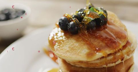 Pouring maple syrup over stack of fluffy pancakes with blueberries and mint, close-up
