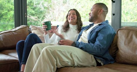 Multicultural couple relaxing on leather sofa drinking coffee by garden window in daylight