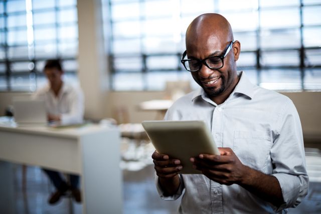 African American businessman using digital tablet in modern office setting. Smiling and engaged, he is focused on the screen. Can be used for business technology, corporate environment, professional workspaces, or digital communication themes.