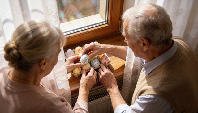 Senior couple arranging pastel easter eggs on sunlit wooden windowsill