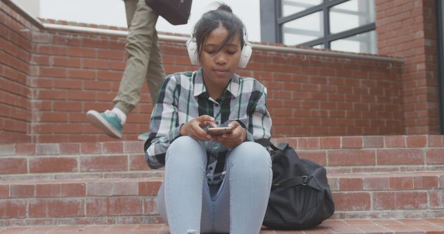 A teenage girl, dressed casually in a plaid shirt and jeans, is sitting on school steps with a brick building in the background, wearing headphones and using a smartphone. The scene exudes a sense of relaxation and focus as she engages with technology. This visual can be used for educational, lifestyle, or youth-oriented content, highlighting themes of youth, technology, education, music, and relaxation.