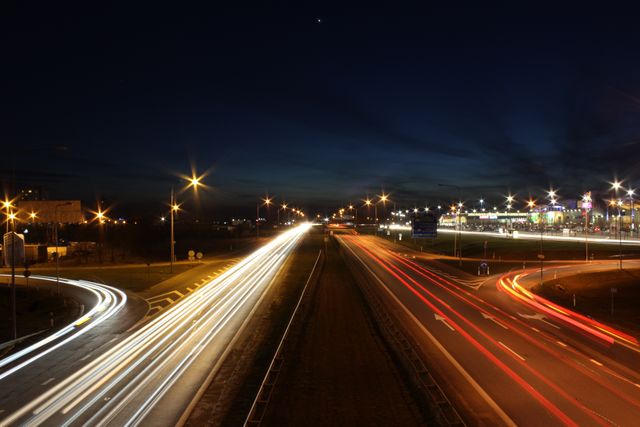 Depicting busy highway with long exposure photograph capturing vivid light trails from moving vehicles. Street lights illuminate road and surrounding area, with dark sky offering contrast to bright lights. Ideal for illustrating concepts of travel, speed, urban life, and transportation. Perfect for articles, blogs on city life or transportation infrastructure, and advertising campaigns for automotive or travel industries.