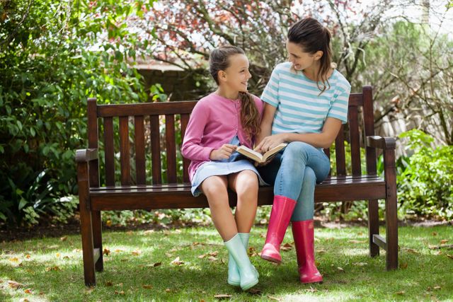 Cheerful mother and daughter reading novel while sitting on wooden bench at backyard