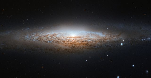 This edge-on view of the UFO Galaxy NGC 2683, captured by the Hubble Space Telescope, showcases the galaxy's delicate dusty lanes silhouetted against its golden core, and scattered clusters of young blue stars. Despite being viewed side-on, astronomers can still deduce the barred spiral structures from the properties of the emitted light. Use this image to illustrate astronomical discoveries, galaxy structures, and the capabilities of Hubble for educational purposes or scientific presentations.