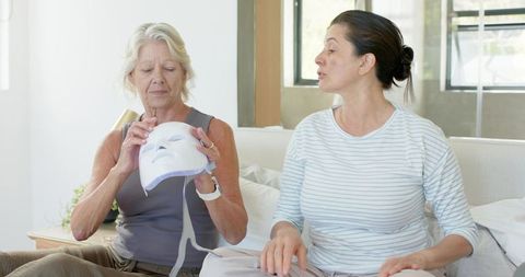 Mature women examining led facial mask together on couch for home skincare and wellness