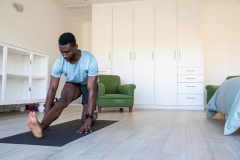 African american man stretching on yoga mat in minimalist sunlit bedroom for home fitness