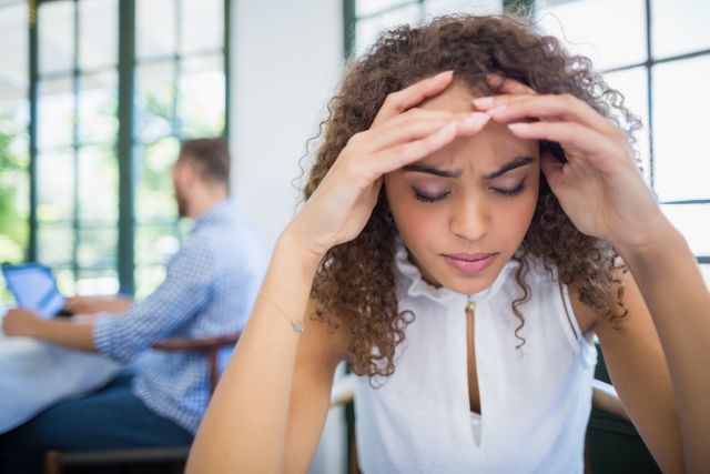 Young woman with curly hair sitting in a restaurant, looking stressed and worried. She is holding her head with both hands, indicating frustration or anxiety. This image can be used to depict stress, mental health issues, or lifestyle challenges in various contexts such as articles, blogs, or advertisements related to mental well-being, stress management, or personal struggles.