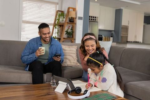 Diverse family watching daughter doing homework at coffee table with tablet and headphones