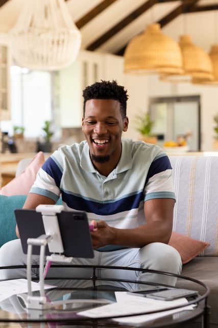 Vertical image of happy african american man using tablet making video call at home, with copy space. Inclusivity, domestic life, leisure time and communication concept.