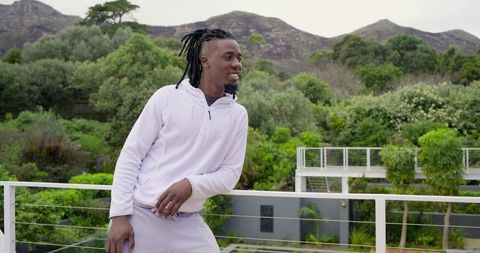 African American Man Leaning on Balcony Wearing Light Hoodie Overlooking Green Terrace
