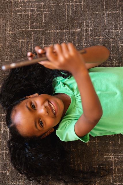 This image shows a young African American girl lying on a carpet and using a smartphone. She is smiling and appears to be enjoying herself. This photo can be used for educational materials, technology advertisements, or articles about childhood and digital learning. It is ideal for promoting apps, online learning platforms, or family-friendly technology products.