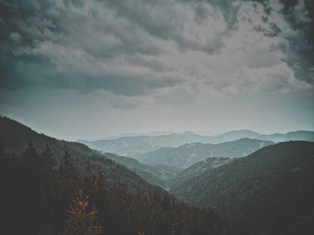 Dark, moody view of a mountain range and forest under an overcast sky. Captures dramatic and serene atmosphere of nature. Ideal for backgrounds, wallpapers, travel blogs, or environmental projects.