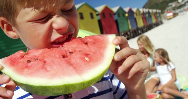 A young Caucasian boy is enjoying a juicy slice of watermelon on a sunny beach, with copy space. Behind him, a group of people, his family, are relaxing on the sand, enhancing the feel of a leisurely summer day.
