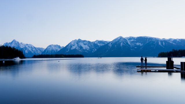A couple stands on a dock, gazing at a serene lake with snow-capped mountains in the background. The calm water reflects the scenic landscape, creating a sense of peace and tranquility. This image can be used in travel promotions, outdoor adventure blogs, meditation or relaxation content, and environmental conservation campaigns.