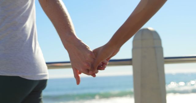 Two people are holding hands by the beach, with copy space. Their interlocked fingers suggest a close relationship or romantic connection against the serene backdrop of the ocean.