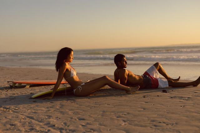 Young couple sitting on sandy beach with surfboards, enjoying sunset. Ideal for travel, vacation, and lifestyle themes. Perfect for promoting beach resorts, surf schools, and summer activities.