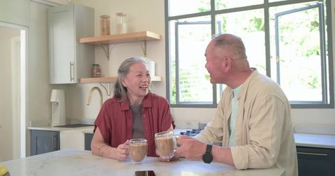Senior couple laughing over coffee at sunlit kitchen island, holding glass lattes