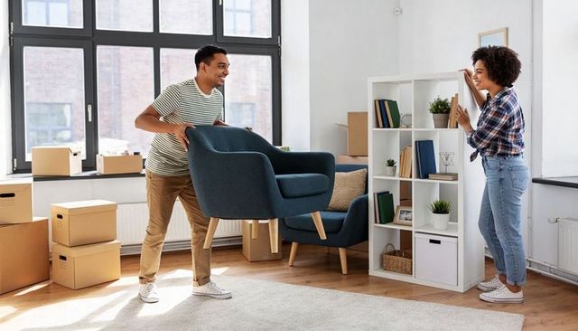 Young couple moving and unpacking in sunlit apartment arranging blue armchair and shelf