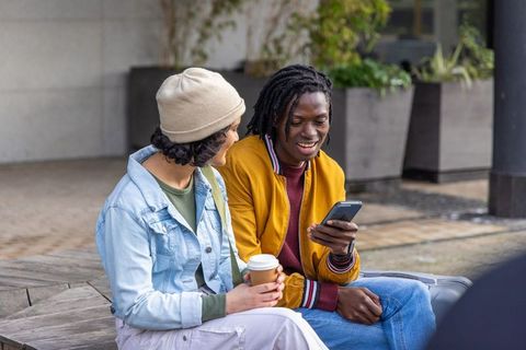 Young diverse friends laughing and sharing smartphone while sipping coffee in urban plaza