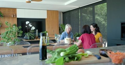 Three Generations Chatting Around Wooden Kitchen Table with Plants, Wine, Fresh Greens