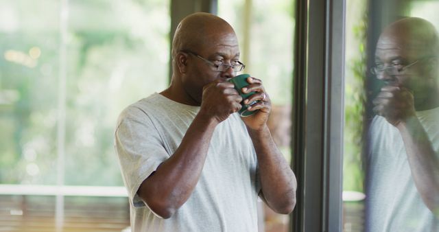 Elderly man holding a green mug and enjoying a hot beverage while standing by a window. Reflection of the man is visible on the glass. Suitable for themes involving relaxation, morning routines, seniors' lifestyle, and peaceful moments.