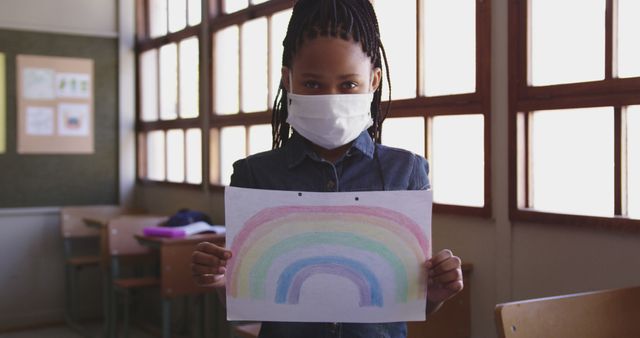 Young girl in school holding up her rainbow drawing while wearing a face mask. Perfect for illustrating creativity in a classroom setting during pandemic, child education in times of COVID-19, or promotion of health and safety measures in schools.