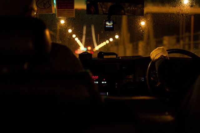 A scene showing a taxi driver navigating urban streets at night with raindrops on the windshield and city lights illuminating the scene. Useful for themes highlighting transportation, city life, or late-night commutes.
