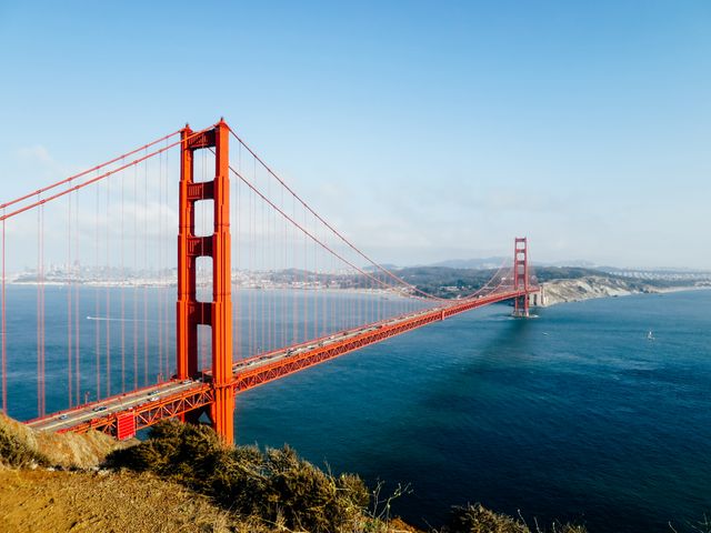 Photo of the Golden Gate Bridge on a clear day with blue sky and calm water. Perfect for articles about travel, tourism, iconic landmarks, and engineering marvels. Ideal for social media posts, travel guides, promotional materials for trips to San Francisco, or educational resources about architectural infrastructure.