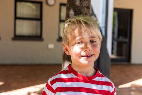 Smiling school-age boy standing on porch wearing red striped shirt under dappled sunlight
