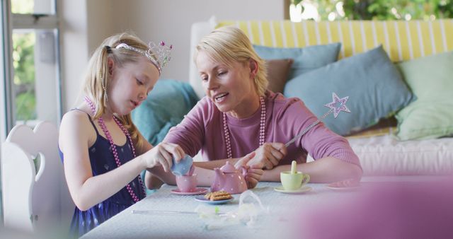 Mother and daughter are engaging in a playful tea party indoors, showcasing their strong bond and happy moment. The girl is dressed as a princess with a crown and beads, adding an imaginative touch. Ideal for advertisements, family-oriented content, and articles on parenting or early childhood development, emphasizing love and togetherness.