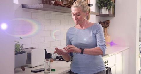 Mature woman taking medication at home counter, preparing daily pill in bright kitchen