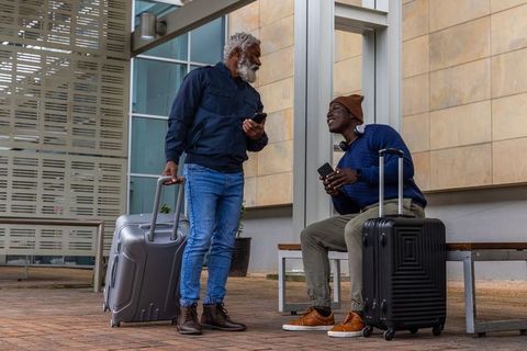 African american men chatting with suitcases at outdoor transit stop, travelers on phones