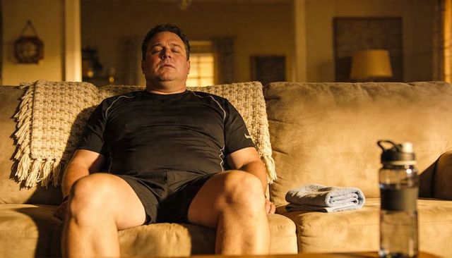 Middle-aged man resting on couch after home workout with towel and water bottle in warm light