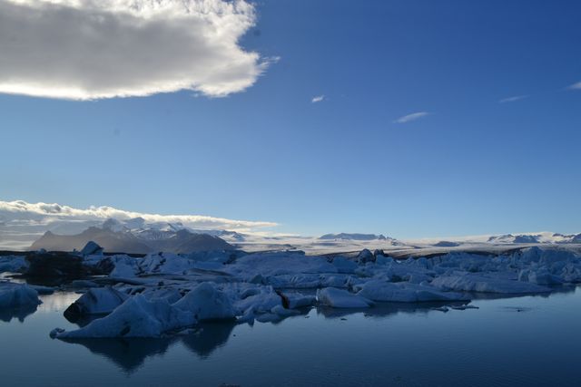 Wide shot showing icy landscape with large icebergs floating on reflective water, set under a clear blue sky. Nearby snow-capped mountains provide a dramatic background. Ideal for use in projects focused on natural beauty, winter scenes, arctic expeditions, climate change, or serene travel destinations.