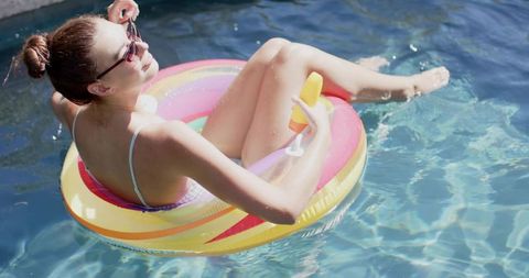 Young woman relaxing on rainbow inflatable float sipping orange drink in sunny pool