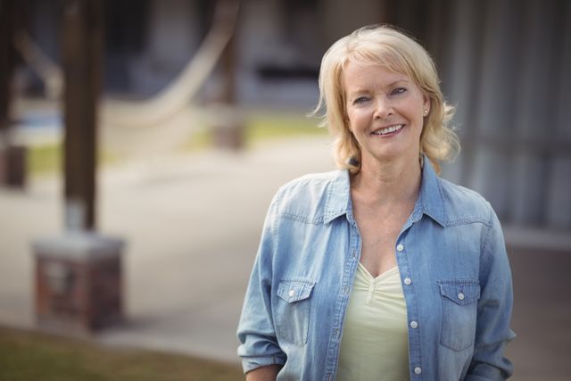 Senior woman smiling outdoors, wearing a denim shirt and standing in a relaxed manner. Ideal for use in lifestyle blogs, retirement community advertisements, health and wellness articles, and promotional materials for senior living.