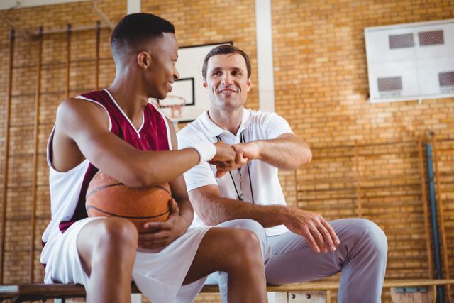 Basketball player doing fist bump with coach while sitting on bench in court