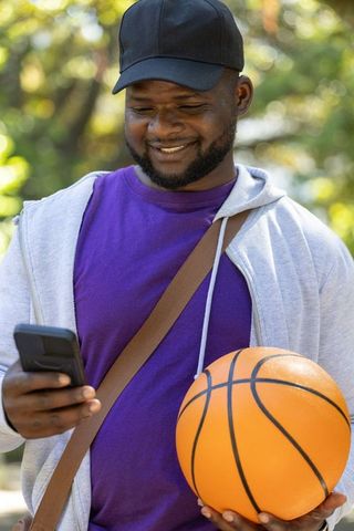 Smiling african american man holding basketball and checking smartphone in sunny park