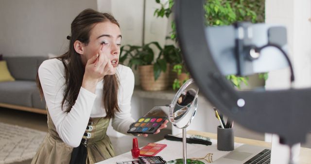 Young woman is applying makeup while recording a tutorial at home. This could be used in articles and posts about beauty, social media influencers, makeup tutorials, and content creation. Ideal for brands targeting beauty enthusiasts, aspiring makeup artists, and social media influencers. Suitable for websites, blogs, online advertising, or use in guides and ebooks about makeup and vlogging.
