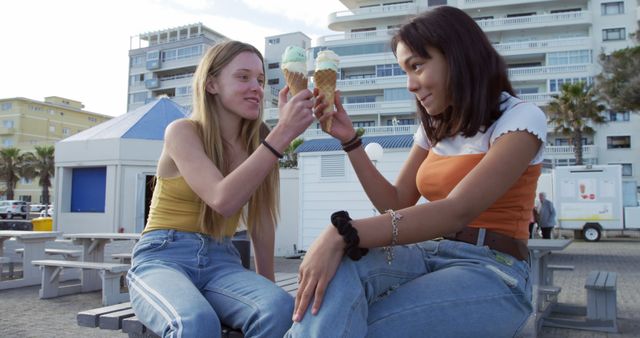 Two young women enjoying ice cream together, sitting on a bench in a beachfront area. The background shows apartment buildings and seaside structures under a clear sky, suggesting a casual and relaxing environment. Perfect for use in advertising summer activities, vacation spots, and highlighting the joy of friendship and leisure.