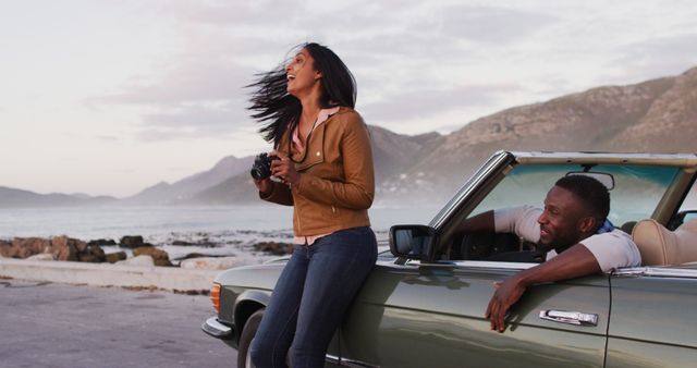 African american woman using digital camera to take pictures while standing near the convertible car on the road. road trip travel and adventure concept