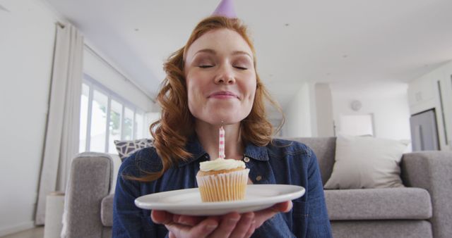 A cheerful woman wearing a party hat smiles with closed eyes, holding a plate with a cupcake and lit candle. The cozy interior of a modern, light-filled home serves as a backdrop. This image is perfect for birthday-related content, celebratory promotions, personal milestones, and cheerful branding.