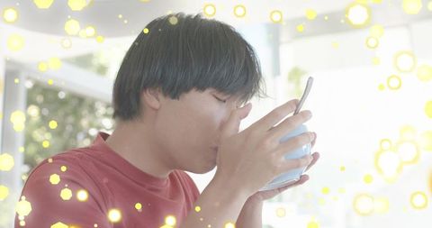 Young asian man sipping from blue mug with spoon during sunlit cozy morning bokeh
