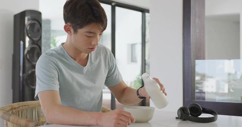 Young man pouring milk into cereal bowl at modern minimalist kitchen table