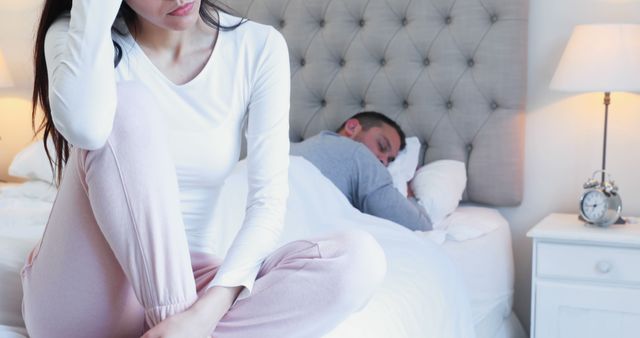Woman sitting on side of bed looking stressed with partner sleeping in background. Possible uses include illustrating relationship problems, mental health articles, blogs about insomnia or anxiety.