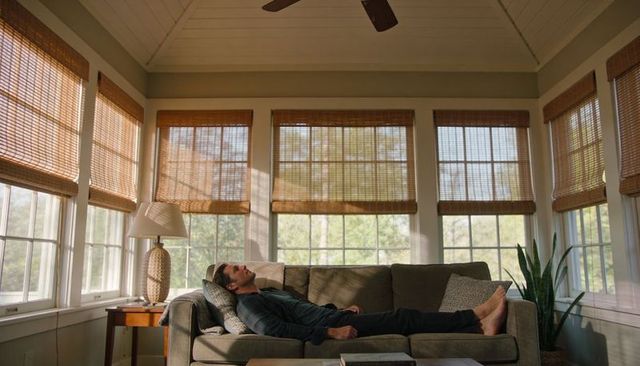 Reclining man resting on sunroom sofa with bamboo blinds, natural light, cozy interior