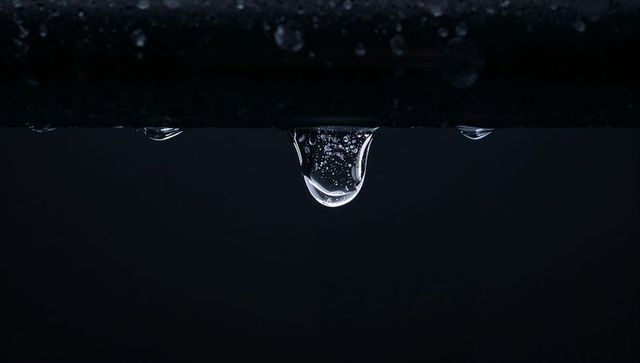 Hanging droplet revealing tiny bubbles and bright reflections on dark metal bar