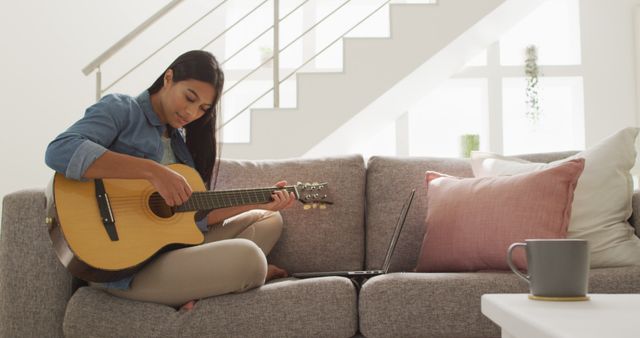 Young woman sitting on a cozy couch in living room, playing an acoustic guitar. Sunlight filters through the window. Ideal for use in content related to home lifestyle, music practice, relaxation, creativity, and casual living.