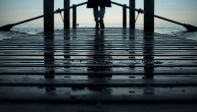 Silhouetted person walking on wet wooden pier at moody overcast seascape with reflections