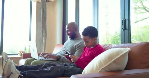 African american father working on laptop while young son playing smartphone on sunlit sofa